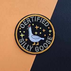 May include: Embroidered patch with a black background and a yellow border. The patch features a white goose with a red crown and the words "CERTIFIED SILLY GOOSE" in yellow. Yellow stars are scattered around the goose.