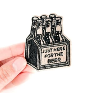 May include: Embroidered patch featuring a black and beige beer bottle carrier with six bottles. The patch has the text "JUST HERE FOR THE BEER" in a rectangular frame. The patch is held by a hand.