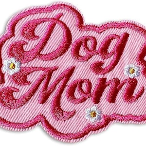 May include: A pink embroidered patch with the words "Dog Mom" in a cursive font. The patch has a scalloped edge and small white flower accents. The text and border are a darker shade of pink.