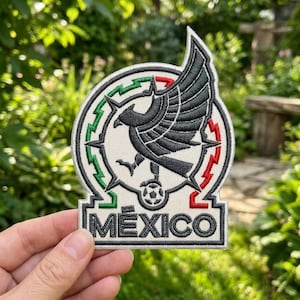 May include: A circular embroidered patch featuring a black eagle with spread wings perched on a soccer ball. The design is framed by green and red lightning bolt patterns on a white background, with the word MEXICO stitched in black at the base.
