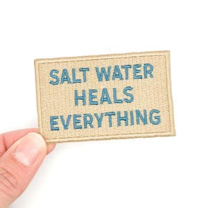 May include: A rectangular patch with the embroidered text "SALT WATER HEALS EVERYTHING" in blue on a tan background. The patch is held by a hand against a white background.