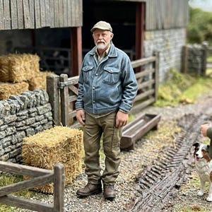 May include: A detailed miniature model of a farmer in a blue jacket and cap, standing near a barn and hay bales. The scene includes a dog and a stone wall, creating a rural setting.