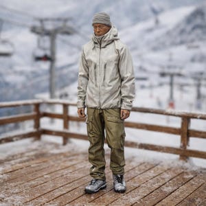 Może przedstawiać: Osoba w jasnoszarej kurtce z kapturem z wieloma kieszeniami zapinanymi na zamek, oliwkowozielonych spodniach cargo, szarej czapce i szarych butach turystycznych. Osoba stoi na drewnianym tarasie pokrytym śniegiem, z wyciągiem narciarskim i ośnieżonymi górami w tle.