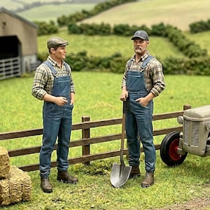 Peut inclure: Deux figurines de fermiers en salopettes en jean et chemises à carreaux se tiennent près d'une clôture en bois. L'un tient une pelle, et un tracteur est partiellement visible. La scène se déroule dans un champ vert avec une grange en arrière-plan.