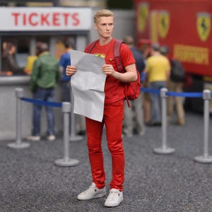 Può includere: Una statuetta di una persona in abiti rossi, che tiene in mano un foglio bianco, con uno zaino rosso e scarpe da ginnastica bianche. La statuetta è in piedi davanti a una biglietteria con la scritta "TICKETS" in lettere rosse. Un camion Ferrari rosso è sullo sfondo.
