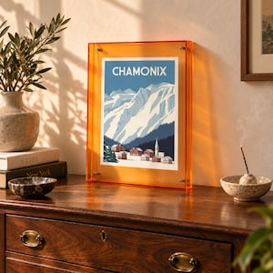 Può includere: Una cornice rettangolare con un bordo in acrilico arancione brillante contenente una stampa di un paesaggio montano innevato. La stampa presenta il testo CHAMONIX in lettere maiuscole bianche sopra montagne blu, pini scuri e un piccolo villaggio.