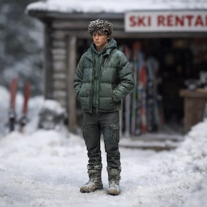 May include: A miniature figure of a person in winter attire stands in front of a ski rental shop. The figure wears a green puffer jacket, a hooded sweatshirt, and dark pants. Snow covers the ground and the figure's hair. The sign above reads "SKI RENTAL."