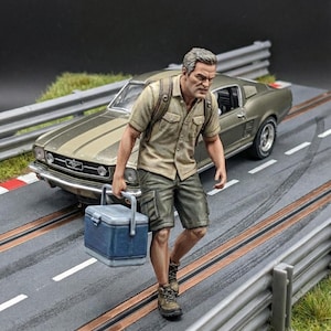 Puede incluir: Una escena de diorama en miniatura que presenta a un hombre que lleva una nevera azul, caminando por una pista de carreras. Un coche Mustang verde oliva clásico está estacionado cerca. El hombre viste camisa color canela, pantalones cortos cargo y botas de montaña.
