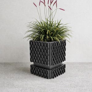 Puede incluir: Una maceta moderna de color gris oscuro con un diseño geométrico, llena de hierba verde y rematada con delicadas espigas de flores moradas. La maceta tiene un patrón texturizado único y se encuentra sobre una superficie gris.