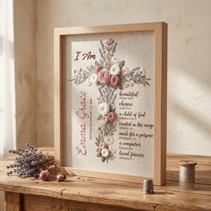 May include: A framed embroidery art piece featuring a cross made of pink and white fabric flowers and green leaves on a beige linen background. The text reads I Am, Emma Grace, Baptized on February 1st, 2026, and various Bible verses.