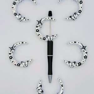 May include: A black pen with silver detailing and a decorative crescent moon design. The moon features black floral and butterfly details. Six matching crescent moon charms with similar designs surround the pen.