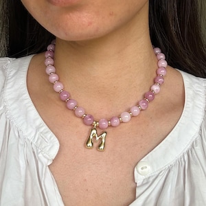 May include: A pink beaded necklace with a gold-coloured letter "M" pendant. The necklace features round, light pink beads with gold-coloured spacers. The pendant is a stylized capital letter "M".