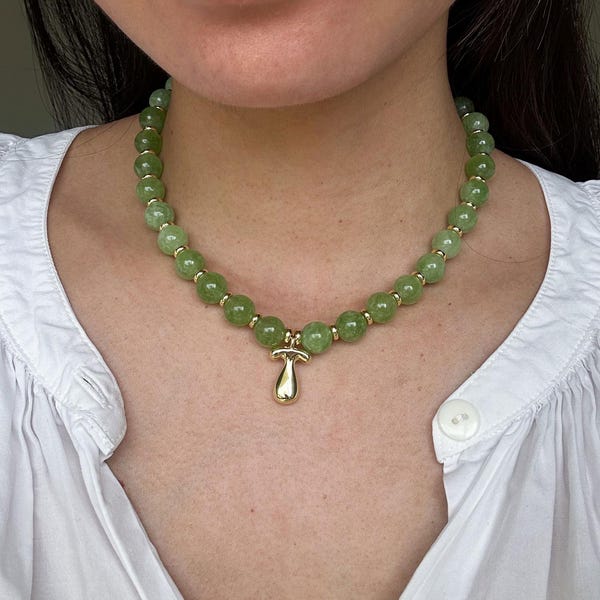 May include: A beaded necklace featuring round, light green beads and gold-colored accents. The necklace has a gold mushroom pendant. The necklace is worn over a white shirt.
