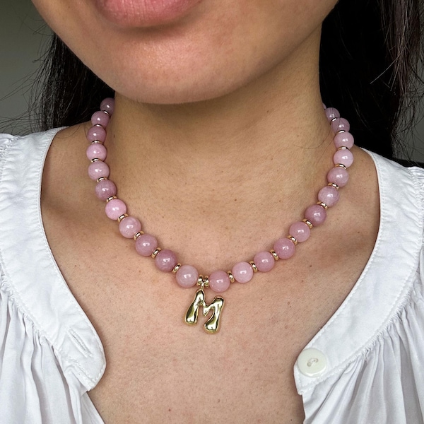 May include: A pink beaded necklace with a gold-colored pendant in the shape of the letter "M". The necklace features round, light pink beads and small gold-colored spacers. The pendant hangs from the center of the necklace.