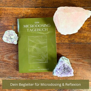 Op de afbeelding: Een groen boek getiteld "DEIN MICRODOSING TAGEBUCH" met een witte lijntekening op de omslag, omringd door kristallen. De tekst "Dein Begleiter für Microdosing & Reflexion" staat onderaan. Het boek ligt op een houten oppervlak.