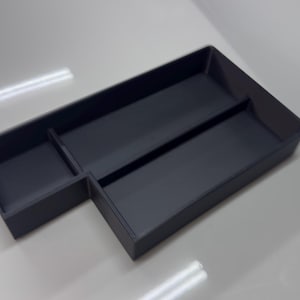Può includere: Un organizer in plastica grigio scuro con più scomparti. L'organizer ha una forma rettangolare con un design sezionato, ideale per riporre piccoli oggetti. La superficie è liscia e riflette la luce.