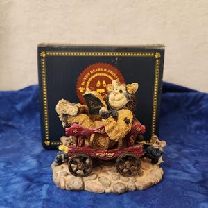 May include: A Boyd's Bears & Friends figurine featuring a bear reading a book while riding in a red wagon. The figurine has a detailed design with a stone base and a dark blue box in the background.
