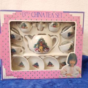 May include: A 13-piece China tea set in a pink box. The set includes a teapot, cups, saucers, and a creamer, all decorated with a Christmas tree and teddy bear design. The box has the words "CHINA TEA SET" printed on it.