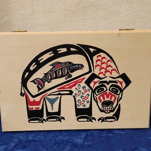 May include: A wooden box with a hinged lid, featuring a stylized bear design in black, red, and blue. The bear has a fish inside its body. The design is reminiscent of Native American art.