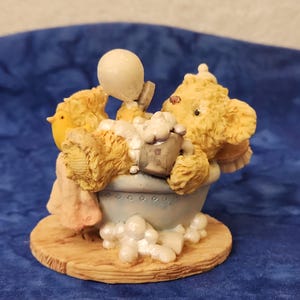 May include: A figurine of a teddy bear and a yellow bird in a bathtub with bubbles. The bear is holding a small bucket and a balloon. The base is a light brown color, and the background is a dark blue.