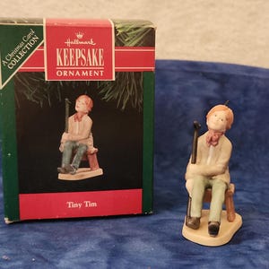 May include: Hallmark Keepsake ornament of Tiny Tim from A Christmas Carol Collection. The figurine is of a young boy with a cane, seated on a small stool. The box is green and red with the Hallmark logo and the words "Keepsake Ornament".