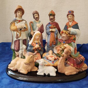 May include: A ceramic nativity scene featuring figures of Mary, Joseph, the Three Wise Men, a shepherd, baby Jesus, and animals. The figures are painted in various colors and stand on a black base. A religious Christmas decoration.