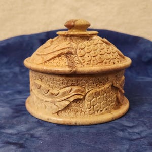 May include: A small, round, beige-colored decorative box with a lid. The box and lid are intricately carved with a repeating pattern of scales and leaves. The lid has a small, rounded knob on top.