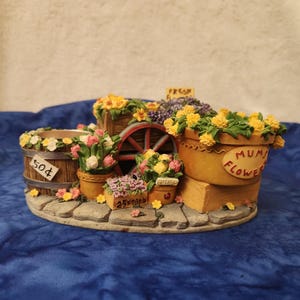 May include: A decorative miniature flower stand with various potted flowers, a wooden wheel, and a sign that reads "Fresh". The stand features a barrel with a "50¢" sign and a pot labeled "Mums Flowers".