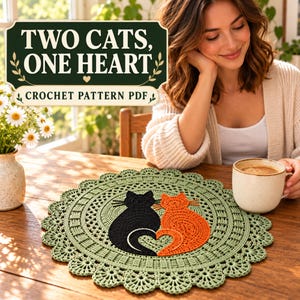 Op de afbeelding: Een rond, lichtgroen gehaakt kleedje met een centraal ontwerp van een zwarte kat en een oranje kat waarvan de staarten een hartvorm vormen. De tekst "TWO CATS, ONE HEART" en "CROCHET PATTERN PDF" staat op een donkergroen rechthoekig bord.