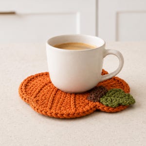 May include: A white ceramic mug filled with dark coffee resting on a round, orange crocheted pumpkin coaster. The coaster features vertical ribbed stitching, a small brown crocheted stem, and two textured green crocheted leaves on the side.