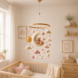 Può includere: Una giostrina sospesa per la cameretta con telaio circolare in legno e figure all'uncinetto, tra cui un orsetto che dorme su una luna crescente, un sole sorridente, nuvole, stelle e piccole sfere nei toni del crema, beige e rosa.