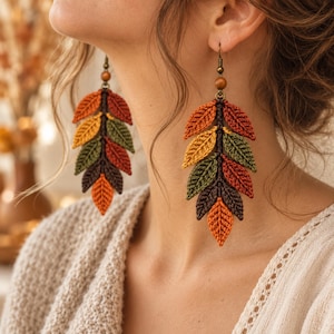 May include: A pair of dangling earrings featuring a vertical arrangement of crocheted leaves in shades of orange, yellow, olive green, and dark brown. Each earring is suspended from a bronze-toned metal hook with a small wooden bead accent.