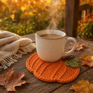 May include: A speckled off-white ceramic mug filled with a steaming hot beverage sits atop a round, orange crocheted pumpkin-shaped coaster. The coaster features a dark brown stem and a small green leaf detail, resting on a weathered wooden surface.