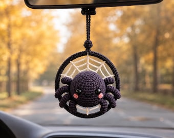 May include: A dark purple crocheted spider ornament hanging from a rearview mirror. The spider is centered within a circular black frame featuring a white crocheted web. It has two large black eyes and small pink stitched cheeks on its round body.