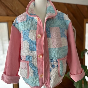 May include: A quilted jacket with a patchwork design in pastel blue, pink, and white. The jacket features pink sleeves, a pink corduroy front, and large, round buttons. The collar and pockets are also trimmed in pink.