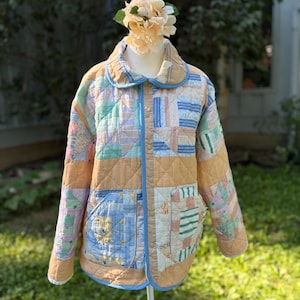 May include: A quilted jacket featuring a patchwork design with various pastel-colored fabric squares in shades of tan, blue, green, and pink. The jacket has a rounded collar, long sleeves, two front patch pockets, and light blue trim along the edges.