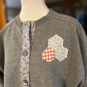May include: A gray sweatshirt with a floral patterned trim and decorative patches. The trim features a light blue floral design, and the patches are a combination of floral and red gingham patterns. The sweatshirt has three gray buttons.