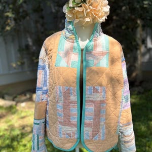 May include: A quilted jacket featuring a patchwork design with tan, light blue, and white fabric panels. The garment has a high collar, long sleeves, and a teal trim along the edges. The fabric displays a mix of solid colors and striped patterns.