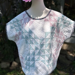 May include: A short-sleeved top constructed from quilted fabric featuring a white base with geometric star patterns in light blue and pink striped fabric. The garment has a round neckline and hem finished with a dark, multi-colored patterned trim.