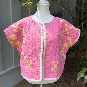 May include: A pink quilted jacket with yellow square patterns and white trim. The jacket is displayed on a mannequin. The short-sleeved jacket has a button-up front and a round neckline. The background includes greenery.