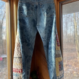 May include: Light blue denim jeans with patchwork quilt fabric accents on the outer seams. The jeans are hanging on a wooden hanger in front of a window. The patchwork includes red, yellow, and blue patterns.