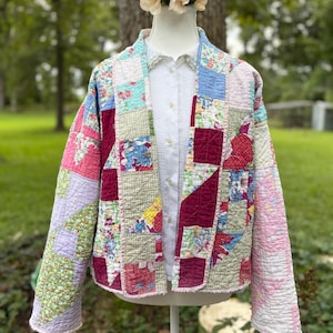 May include: A long-sleeved jacket constructed from a patchwork quilt featuring various floral and geometric fabric patterns in shades of pink, burgundy, light blue, green, and yellow, worn over a white button-down shirt with a lace collar.