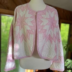 May include: A quilted jacket in white and pink, featuring a starburst pattern. The jacket has long sleeves and a round neckline. The edges are trimmed with a pink polka dot fabric. The jacket is displayed on a mannequin.