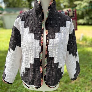 May include: A quilted jacket featuring a geometric log cabin pattern in black and white. The fabric has a subtle textured finish with small floral accents. The interior lining and front edges are trimmed with a contrasting floral print fabric.