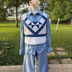 May include: A cropped quilted vest featuring a white and navy blue star-patterned fabric design, worn over a light blue denim button-down shirt and matching light blue denim jeans. The vest has a wide scoop neckline and a structured, sleeveless silhouette.