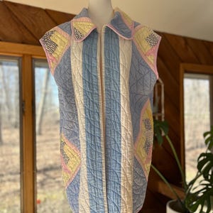 May include: A sleeveless quilted vest in pastel colors, displayed on a mannequin. The vest features a mix of blue, white, yellow, and pink patches, with a vertical striped pattern and a zip-up front. The collar and edges are trimmed with pink fabric.