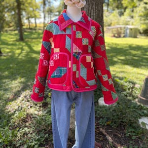 May include: A quilted jacket featuring a patchwork design of red, blue, green, and yellow fabric squares with a bright pink trim along the collar, cuffs, and pockets. The jacket is paired with light blue denim jeans on a display mannequin.