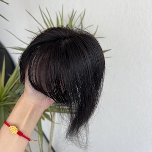 Könnte beinhalten: Ein Haarteil aus glattem, tiefschwarzem Haar mit einem natürlich wirkenden Scheitel und fransigem Pony, der die Stirnpartie umrahmt. Das Haar ist geschmeidig und weist eine gleichmäßige dunkle Farbe auf, um Volumen und Abdeckung zu bieten.