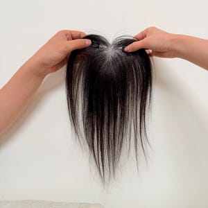 以下が含まれることがあります： 明るい色のベースが付いた、濃い色の髪の毛のヘアピース。ヘアピースは白い背景に対して両手で持たれています。ヘアピースのベースにはクリップが付いています。