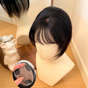 Könnte beinhalten: Ein schwarzes Haarteil mit Pony wird auf einem Schaufensterkopf gezeigt. Das Haarteil ist dunkel und scheint aus Naturhaar zu bestehen. Das Haarteil hat Clips zur Befestigung. Die Schaufensterpuppe steht auf einer Holzoberfläche.